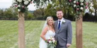 A bride and groom standing under a floral arch in a grassy field.