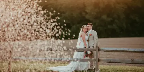 A bride and groom standing together by a wooden fence with confetti falling in the foreground.