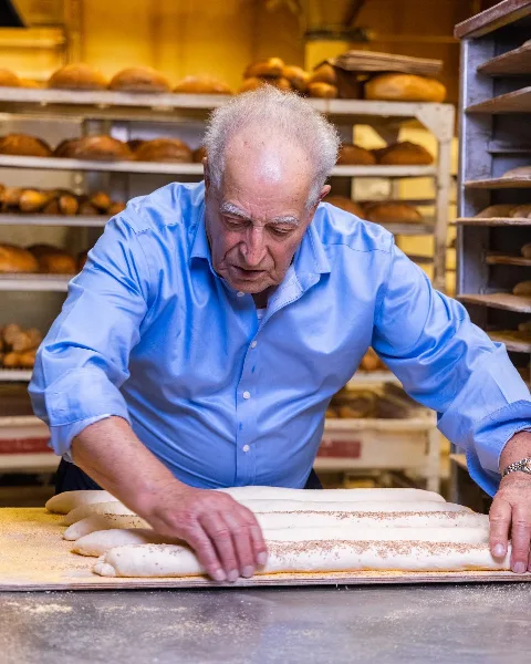 A baker in a blue shirt shaping long loaves of bread on a floured surface in a bakery.