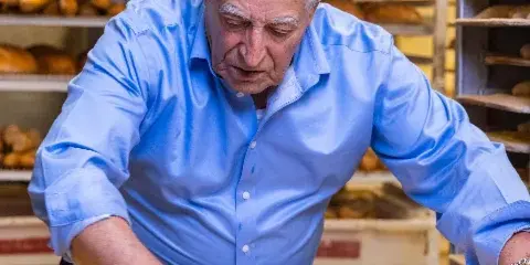 A baker in a blue shirt shaping long loaves of bread on a floured surface in a bakery.