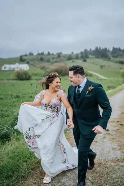 A bride and groom walking hand-in-hand on a dirt path in a grassy field, with the bride holding her floral dress.