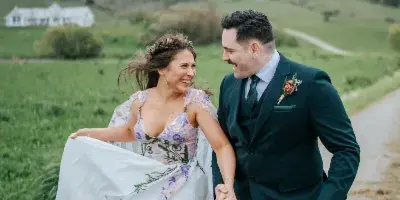 A bride and groom walking hand-in-hand on a dirt path in a grassy field, with the bride holding her floral dress.