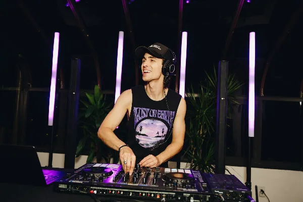 A professional DJ wearing headphones and a cap, operating a Pioneer DJ mixer and turntables in a venue with purple lighting.