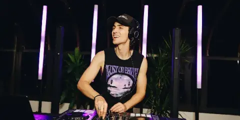 A professional DJ wearing headphones and a cap, operating a Pioneer DJ mixer and turntables in a venue with purple lighting.