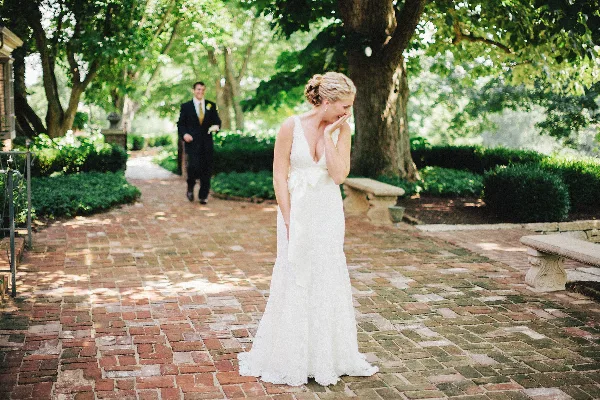 A bride in a white lace gown stands on a brick pathway in a garden, smiling as a groom in a dark suit approaches in the background.