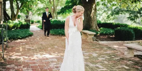 A bride in a white lace gown stands on a brick pathway in a garden, smiling as a groom in a dark suit approaches in the background.