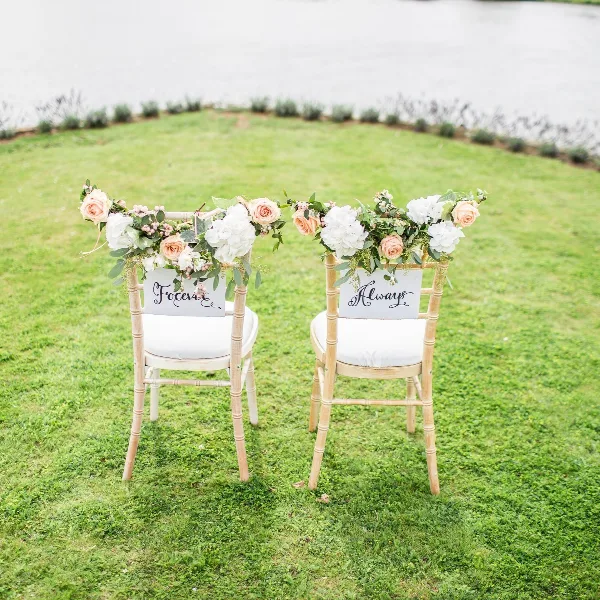 Two white chairs decorated with floral arrangements and signs reading 'Forever' and 'Always' on a green lawn near a body of water.