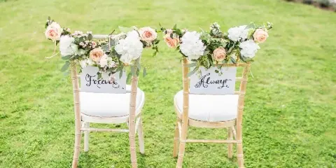 Two white chairs decorated with floral arrangements and signs reading 'Forever' and 'Always' on a green lawn near a body of water.