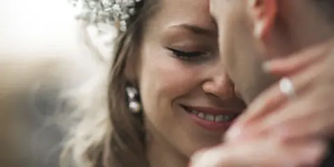 A close-up photograph of a smiling bride with a floral headpiece and a groom in an intimate embrace.