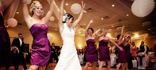 A bride and bridesmaids in purple dresses dancing at a wedding reception.