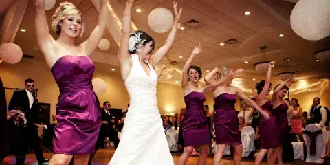 A bride and bridesmaids in purple dresses dancing at a wedding reception.