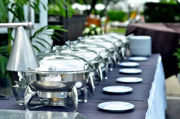 A long buffet table set with a row of stainless steel chafing dishes and white plates in an outdoor event setting.