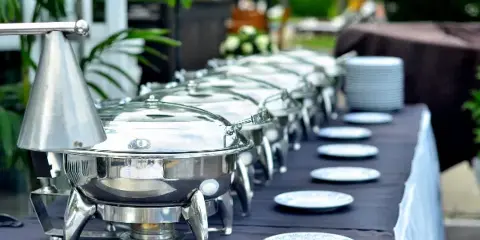 A long buffet table set with a row of stainless steel chafing dishes and white plates in an outdoor event setting.