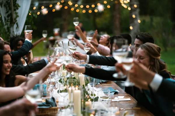 Guests at a wedding reception raising wine glasses in a toast at a long wooden table.