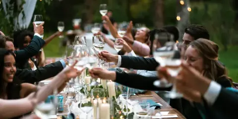 Guests at a wedding reception raising wine glasses in a toast at a long wooden table.