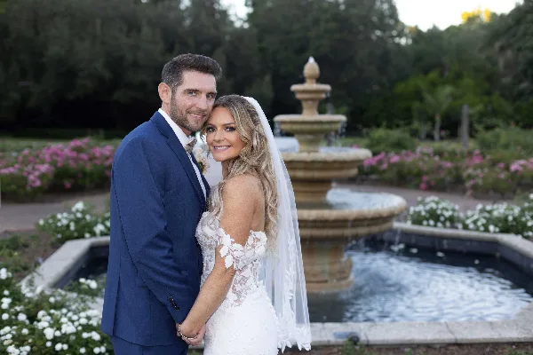 A bride and groom posing together in a garden setting with a stone fountain in the background.