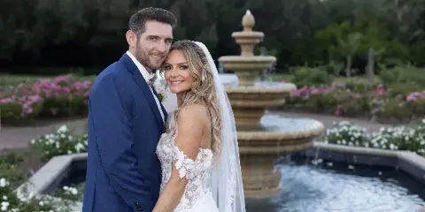 A bride and groom posing together in a garden setting with a stone fountain in the background.