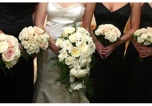 A bride and bridesmaids in black dresses holding white and pink floral bouquets.