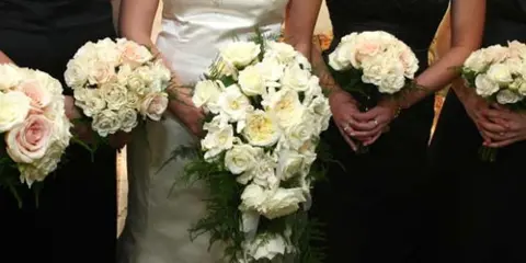 A bride and bridesmaids in black dresses holding white and pink floral bouquets.