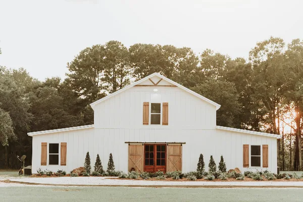 Exterior view of a white barn-style wedding venue with wooden shutters and sliding barn doors, set against a backdrop of trees.
