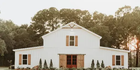 Exterior view of a white barn-style wedding venue with wooden shutters and sliding barn doors, set against a backdrop of trees.