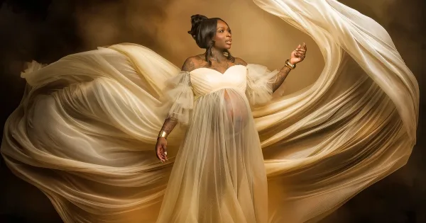 Fine-art maternity portrait of a woman in a flowing white gown with dramatic fabric movement.