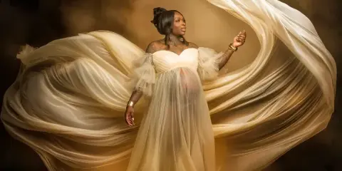 Fine-art maternity portrait of a woman in a flowing white gown with dramatic fabric movement.