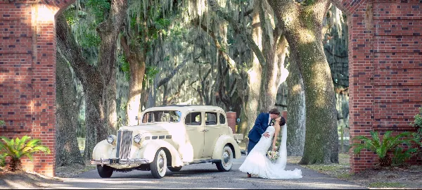 A bride and groom kissing next to a vintage cream-colored car on a tree-lined path with Spanish moss and a brick archway.