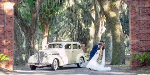 A bride and groom kissing next to a vintage cream-colored car on a tree-lined path with Spanish moss and a brick archway.