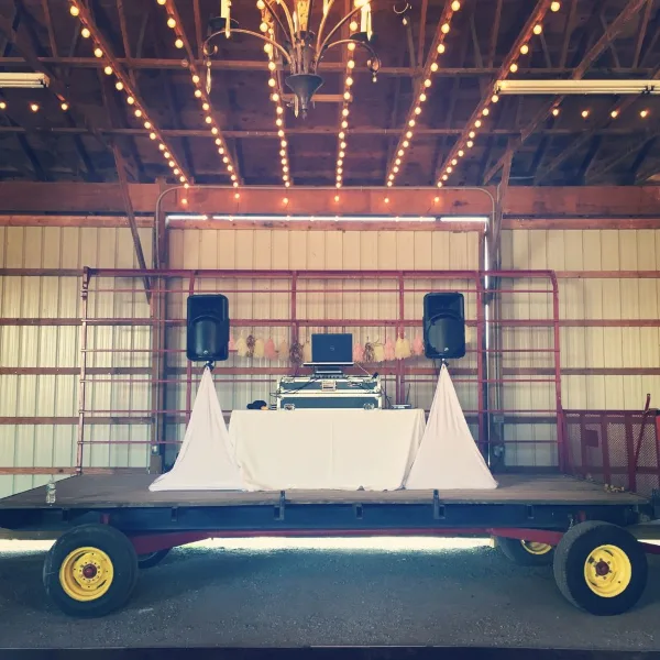 A custom DJ booth and stage setup featuring a table, laptop, and speakers on a wheeled platform in a rustic venue.