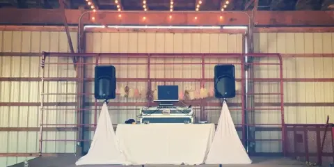 A custom DJ booth and stage setup featuring a table, laptop, and speakers on a wheeled platform in a rustic venue.