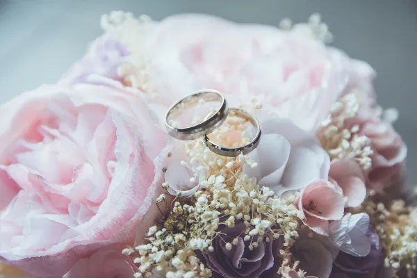 A close-up photograph of two wedding rings resting on a bouquet of pink and white flowers.