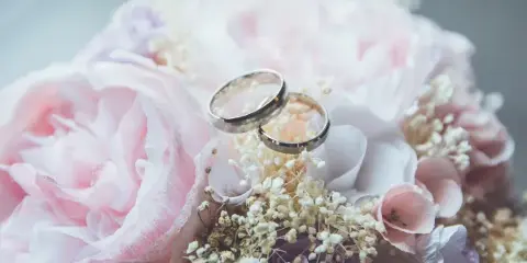 A close-up photograph of two wedding rings resting on a bouquet of pink and white flowers.