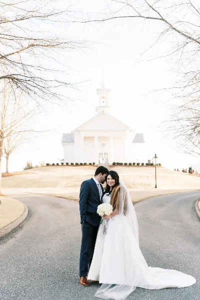A wedding couple standing on a driveway with the white chapel building and bell tower in the background.