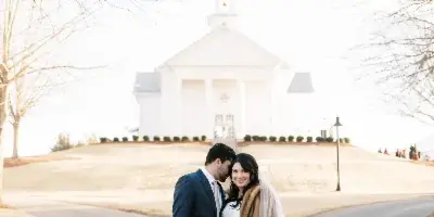 A wedding couple standing on a driveway with the white chapel building and bell tower in the background.