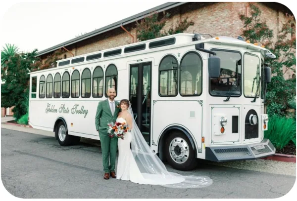 A bride and groom standing in front of a white shuttle bus at a wedding venue.