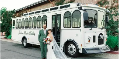 A bride and groom standing in front of a white shuttle bus at a wedding venue.