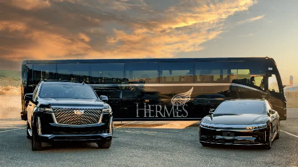 A black Hermes Worldwide coach, a black Cadillac SUV, and a black luxury sedan parked in a row at sunset.