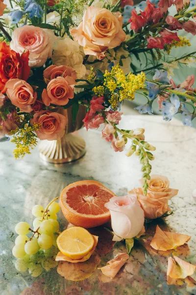 A floral arrangement with peach roses, blue delphiniums, and yellow filler flowers in a gold vase, accompanied by fresh fruit on a marble table.