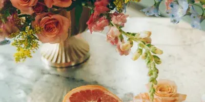A floral arrangement with peach roses, blue delphiniums, and yellow filler flowers in a gold vase, accompanied by fresh fruit on a marble table.