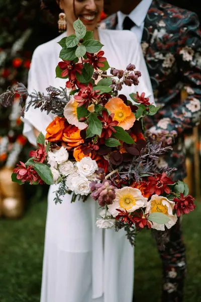 A bride in a white dress holds a large, vibrant bouquet of orange, red, and white flowers with greenery.