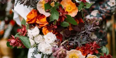 A bride in a white dress holds a large, vibrant bouquet of orange, red, and white flowers with greenery.