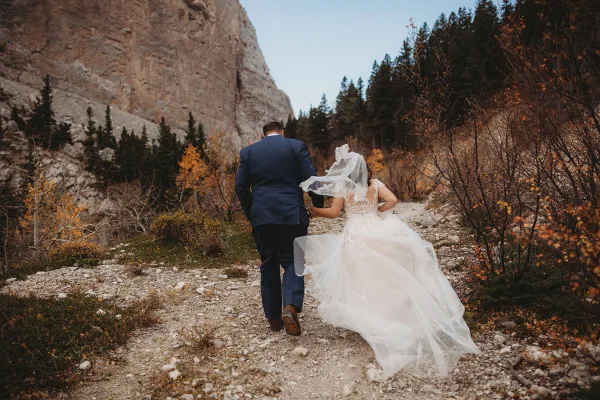 A bride and groom walking hand-in-hand on a rocky mountain trail surrounded by autumn foliage and evergreen trees.