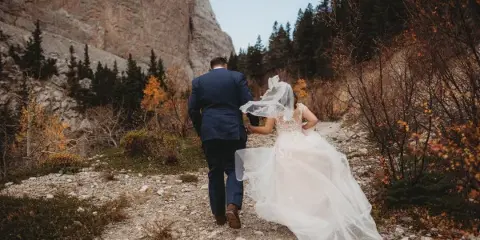 A bride and groom walking hand-in-hand on a rocky mountain trail surrounded by autumn foliage and evergreen trees.
