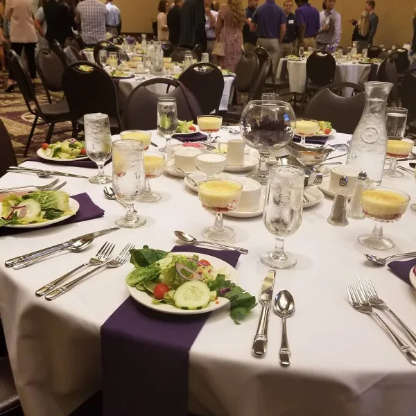 A formal catering table setting featuring a salad, dessert cups, and silverware on a white tablecloth with a purple runner.