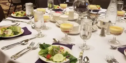 A formal catering table setting featuring a salad, dessert cups, and silverware on a white tablecloth with a purple runner.