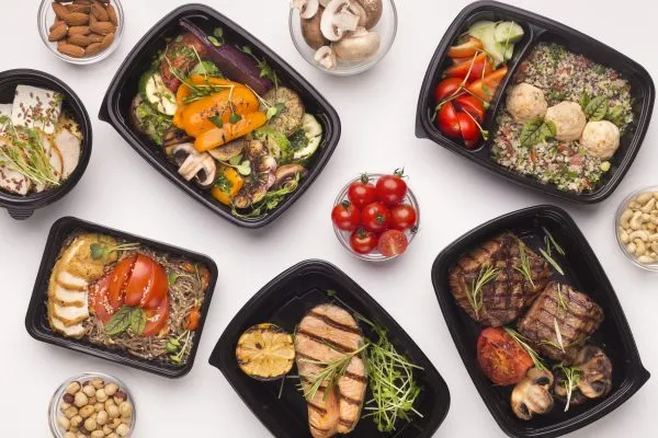 An overhead view of several black meal boxes containing various prepared dishes including grilled chicken, steak, and vegetable medleys, arranged on a white surface.