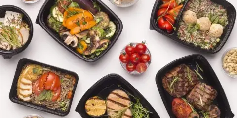 An overhead view of several black meal boxes containing various prepared dishes including grilled chicken, steak, and vegetable medleys, arranged on a white surface.