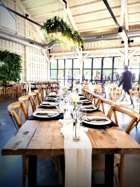 A long wooden dining table set with white plates, black napkins, and glassware in a rustic event venue.