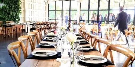 A long wooden dining table set with white plates, black napkins, and glassware in a rustic event venue.